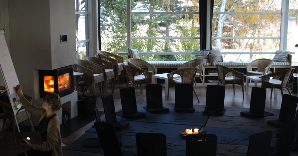 Gathering hall set for a session, low chairs in a circle around lit candles, fireplace and autumn trees beyond the windows