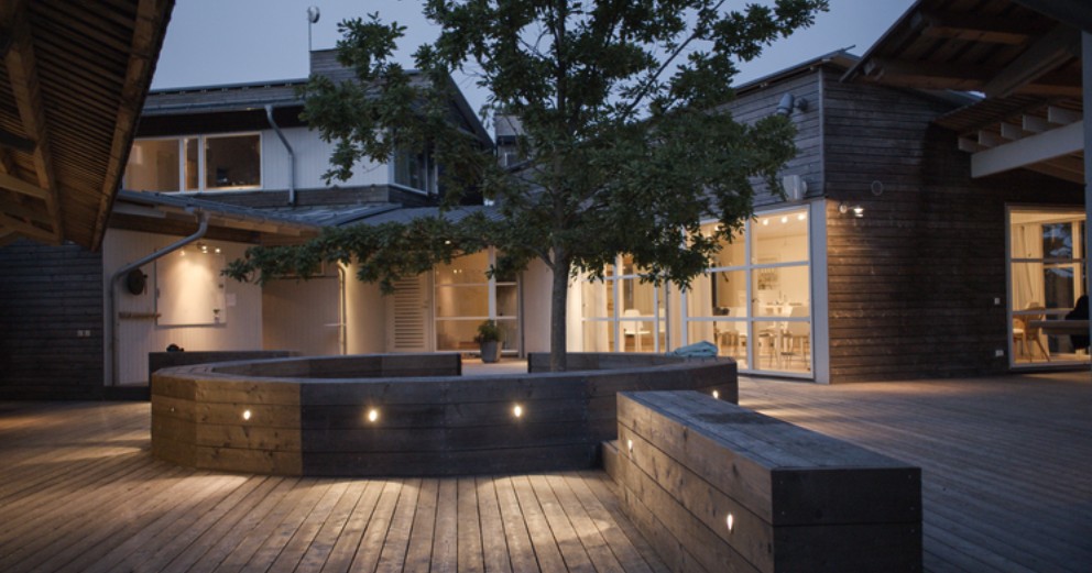 Wooden deck courtyard at dusk, warm lights glowing from the windows and a tree in the centre